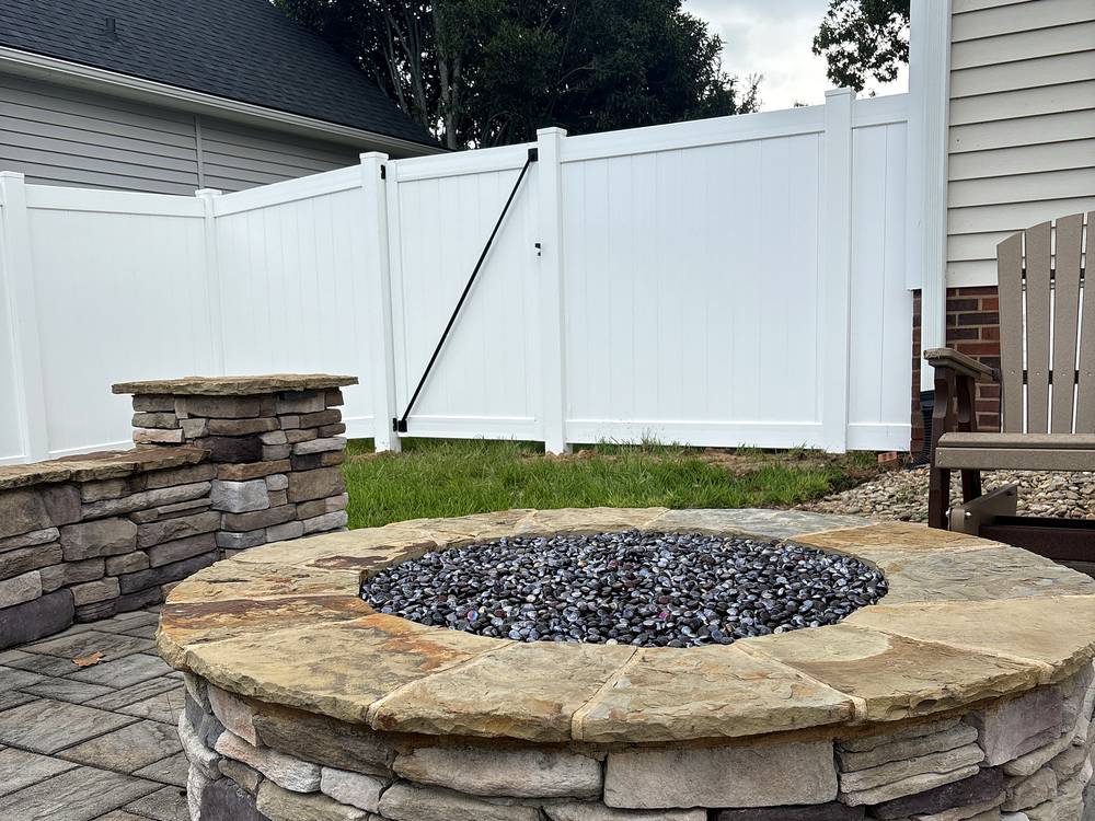 Stone fire pit with a seat wall beside a white vinyl privacy fence in Lexington, SC